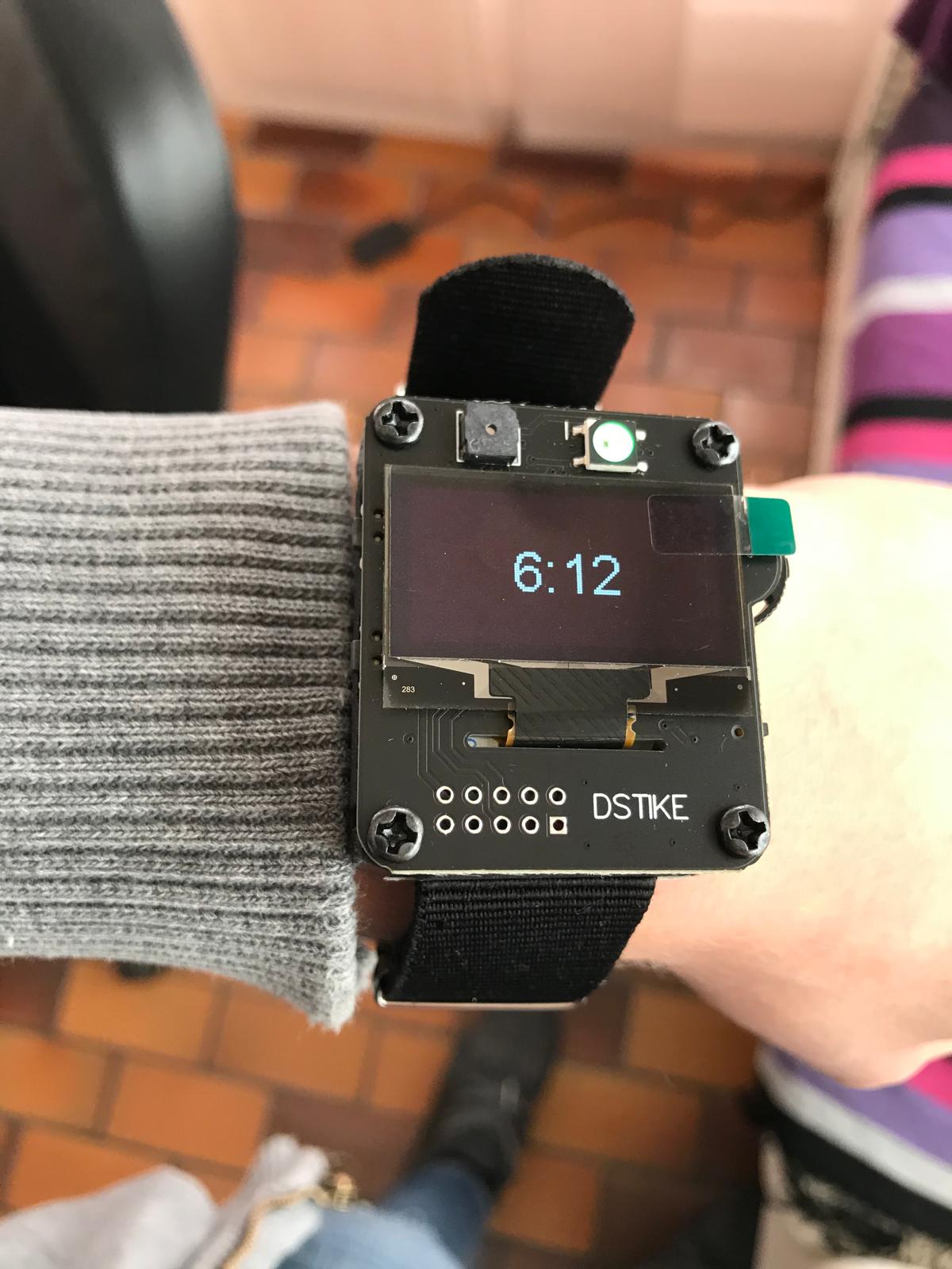 The DSTIKE programmable smartwatch on a wrist — a bare-board watch with an OLED screen reading 6:12, an RGB LED, and the DSTIKE label — used as the remote controller for claiming a device.
