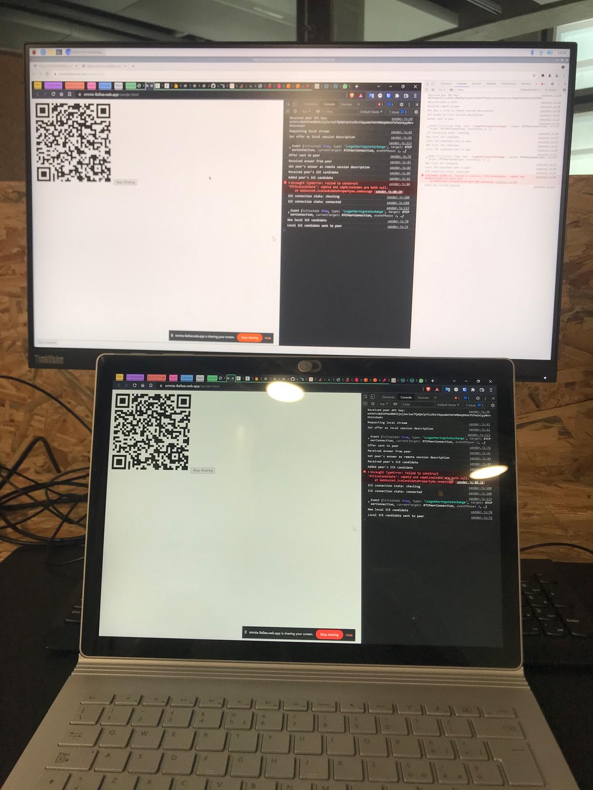A monitor and a laptop side by side, both running the Omnia Share sender page with a QR code and the WebRTC debug console open — screen-sharing between the two.