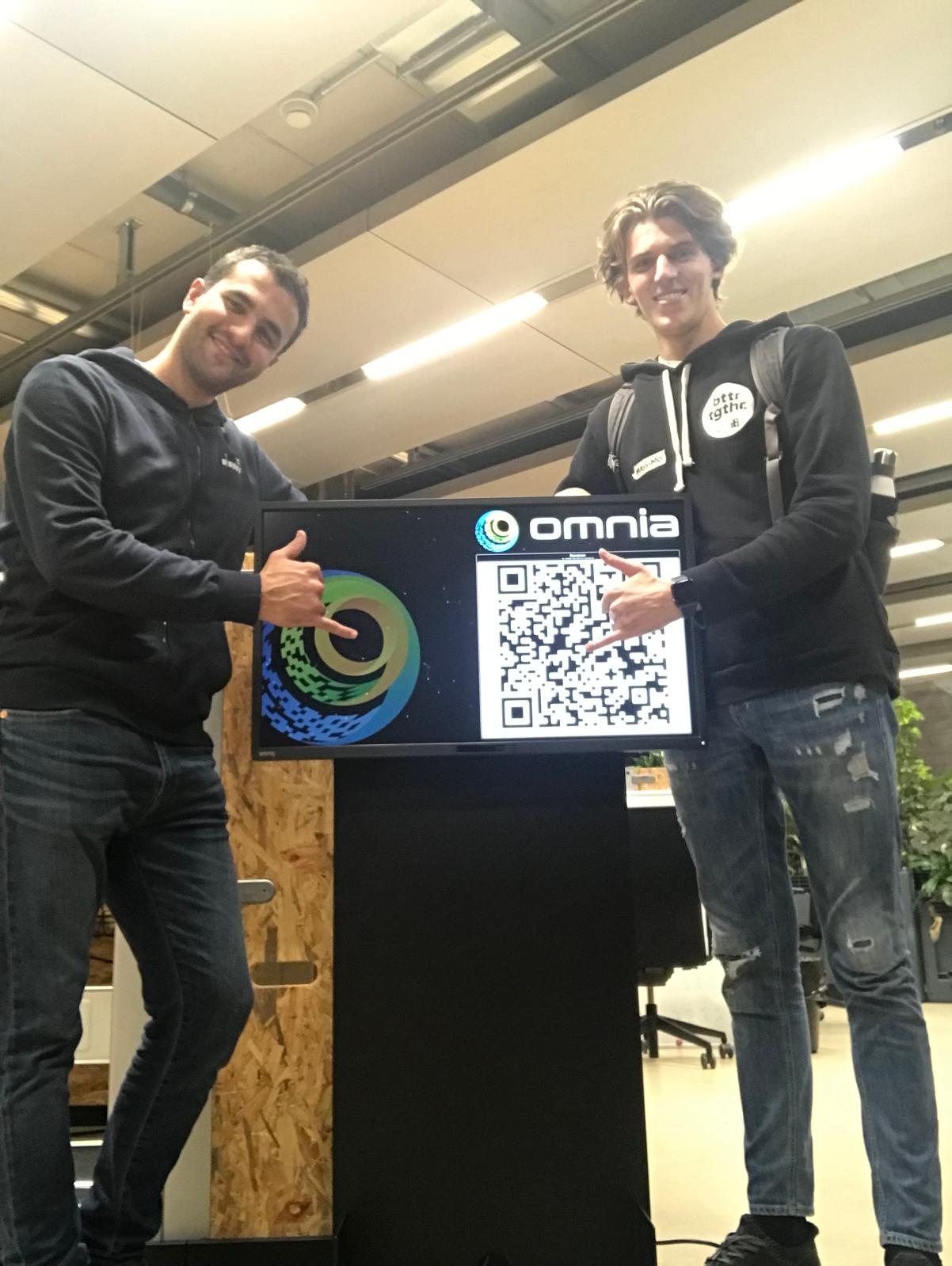 Massimo and a teammate throwing shakas next to a screen showing the Omnia logo and an Omnia Share QR code at SPH.