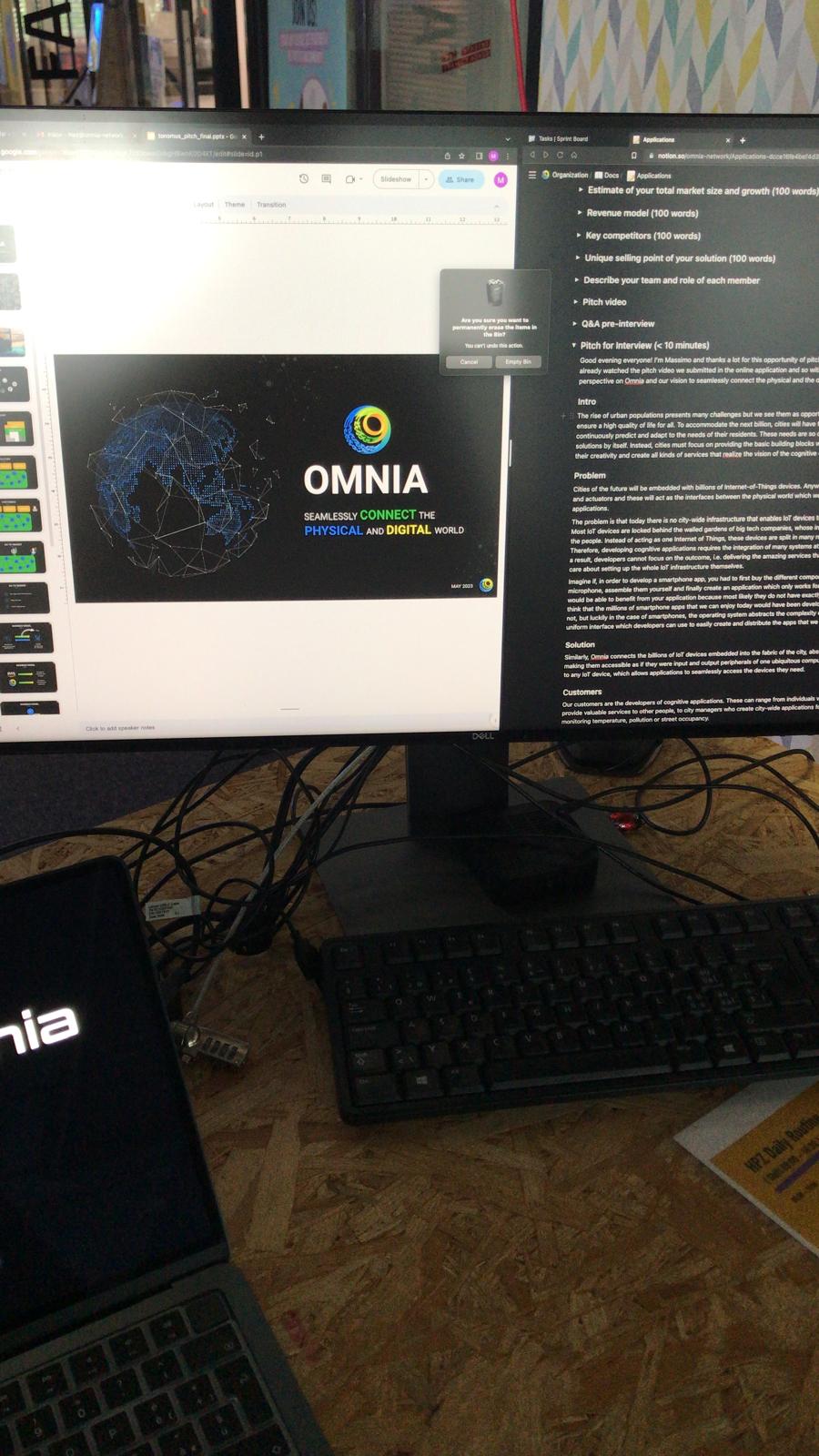 Photo of a desk with a Dell monitor showing the OMNIA pitch-deck title slide ("Seamlessly connect the physical and digital world") in Google Slides next to a Notion applications doc, with an Omnia-branded laptop in the foreground.